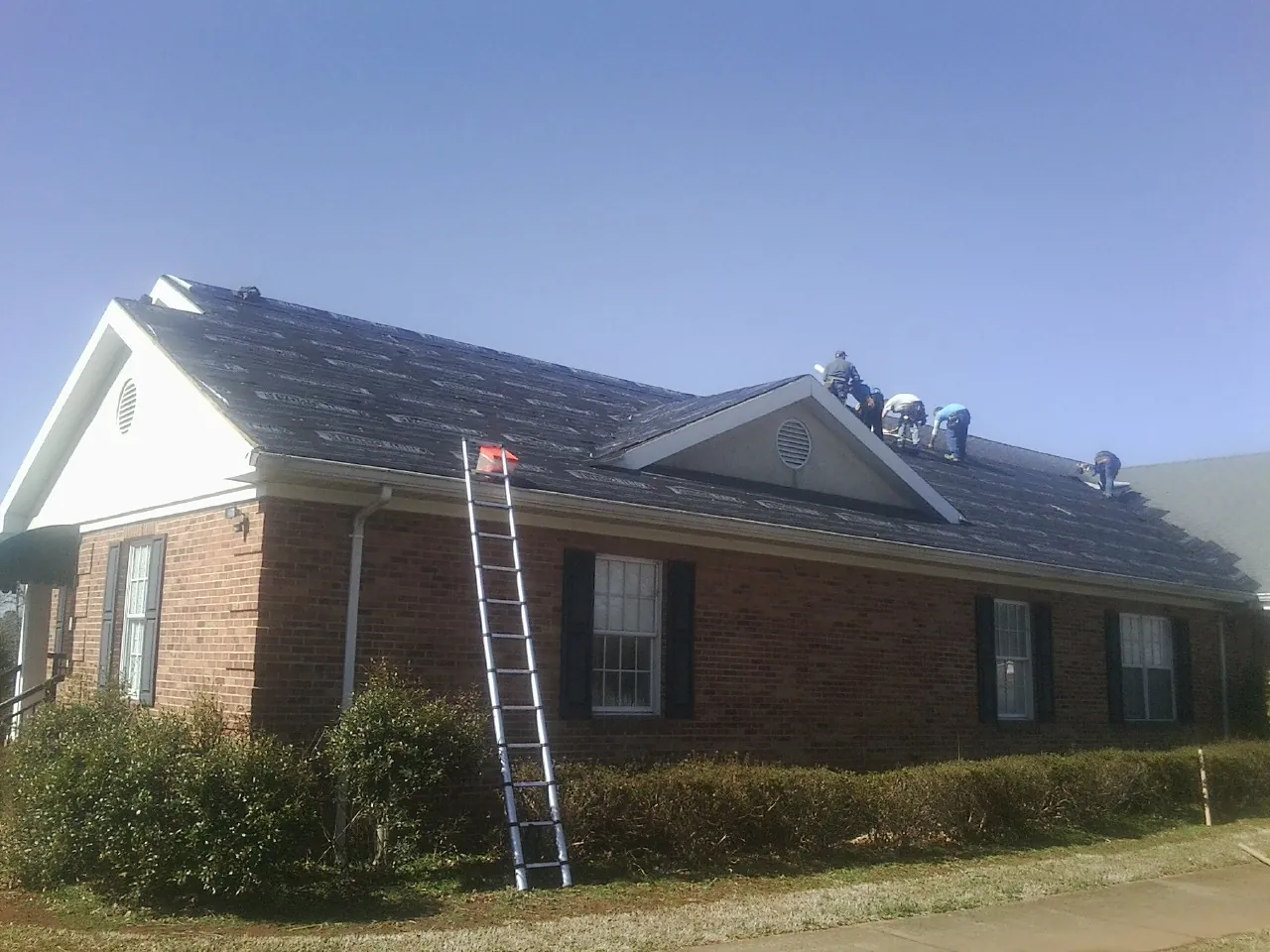 Crew tearing shingles off a brick ranch roof with a ladder set — Emergency Roof Repair work by our crew in Eighty Eight