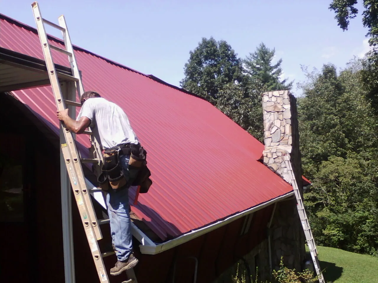 Roofer on a ladder beside a red metal roof and stone chimney — Soffit & Fascia Repair by Roofing Brothers in Eighty Eight